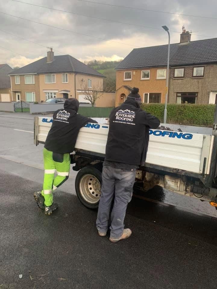 Two roofers in branded jackets leaning into the back of the Riverside Roofing tipper on a street of pebbledashed semis, with fells behind the houses.