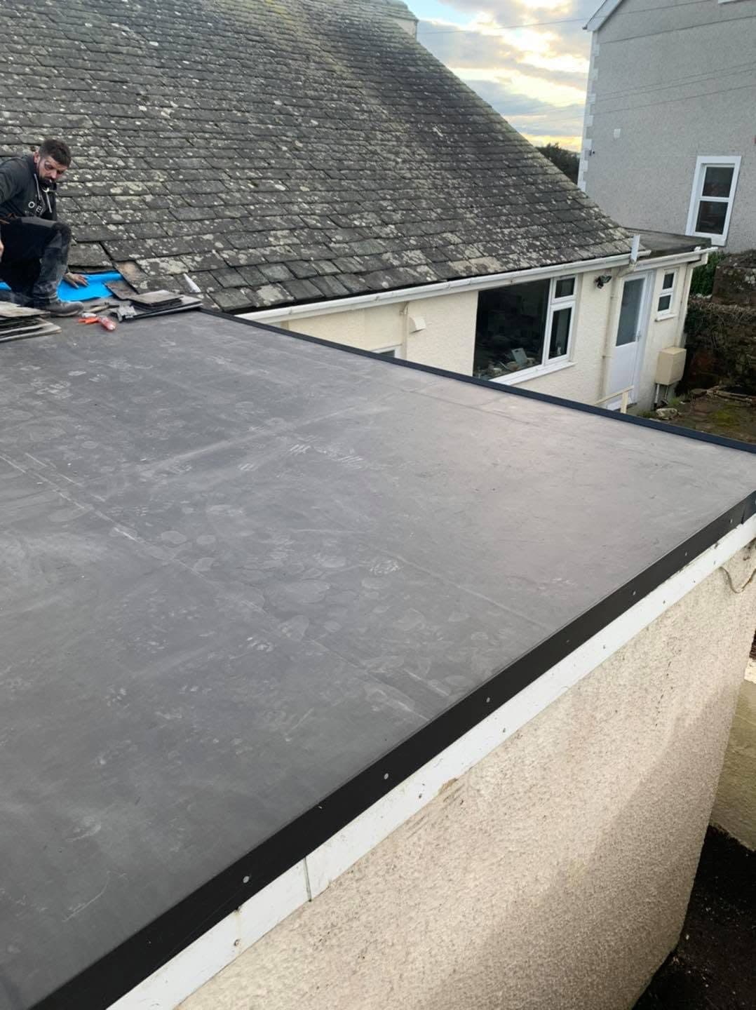 A finished rubber flat roof on a rear extension, laid in one sheet with a black trim along the parapet, the old slate roof of the house behind it.