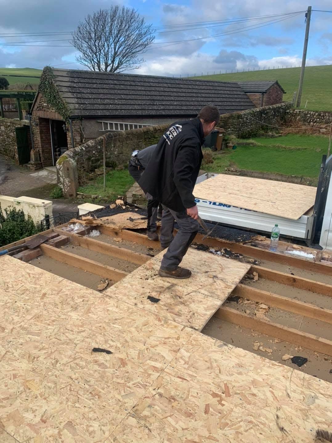 A roofer cutting away rotten boards on a flat roof and laying fresh OSB decking over the joists, with new sheets stacked on the van in the yard below.