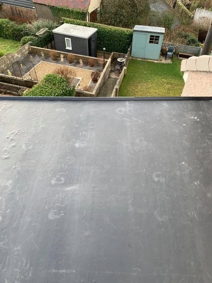 A finished rubber flat roof seen from above, laid in one unbroken sheet to the edge trim, with the garden and sheds below.
