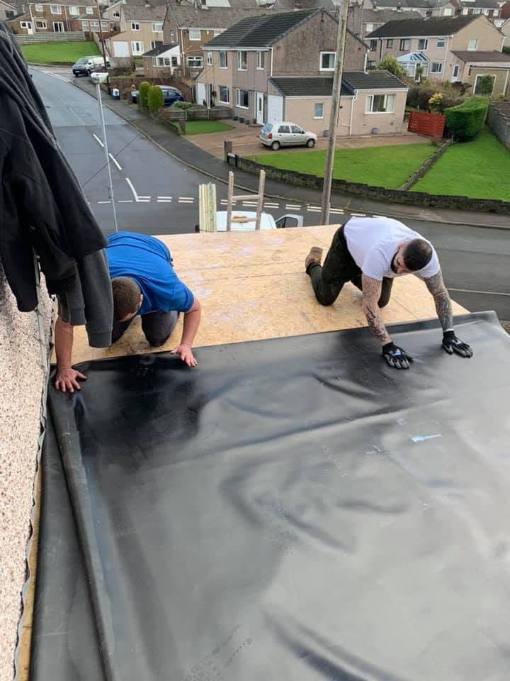 Two roofers rolling a sheet of EPDM rubber out flat across new boards on an extension roof, with the street and houses opposite behind them.
