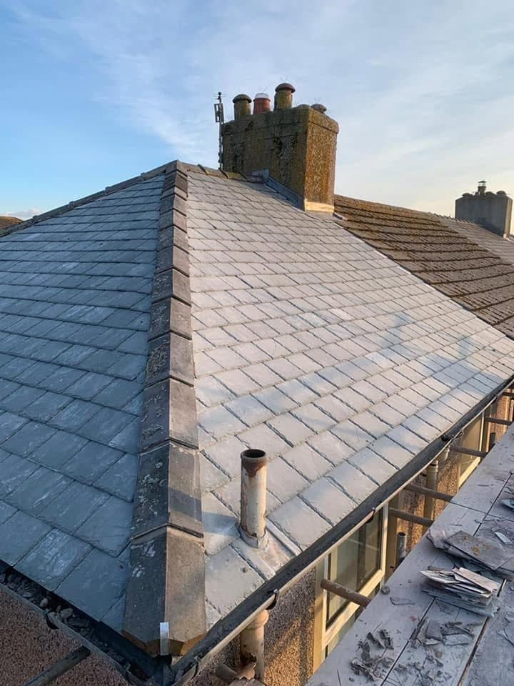 A finished hipped roof in new slate with lead-dressed hips and ridge, the old stone chimney stack standing above it against a clear sky.