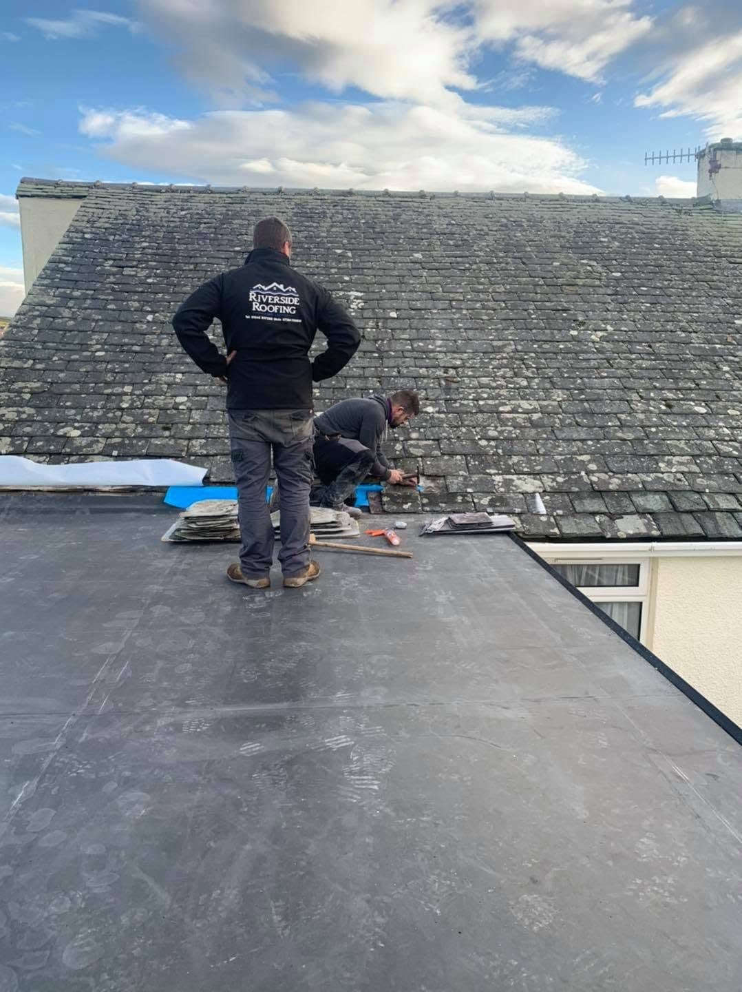 Two roofers standing on a newly laid rubber flat roof, refitting slates on the pitched roof where it meets the extension.