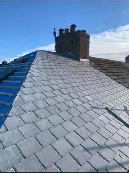 Slating part way up a roof, the new courses laid to the chimney and blue breather membrane still showing on the side yet to be covered.