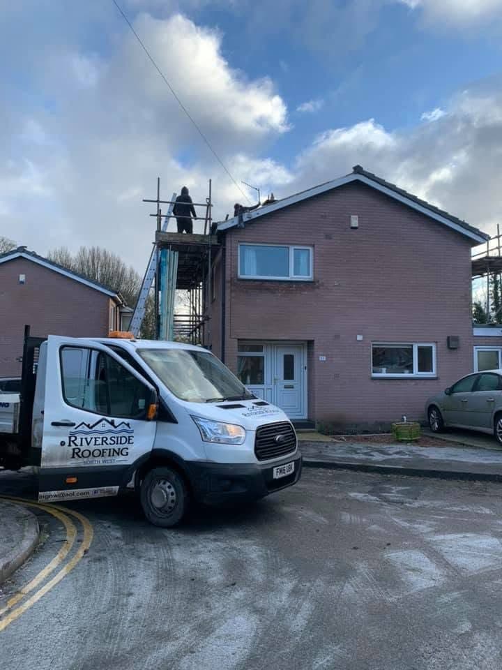 The Riverside Roofing tipper parked outside a brick semi with a tower scaffold up against the gable and a roofer working at the eaves.